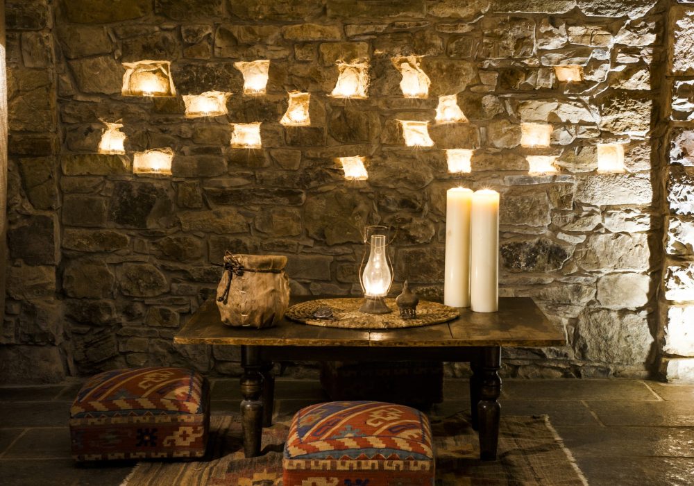 Parete in pietra con nicchie di candele, tavolo con lume a olio e brocca